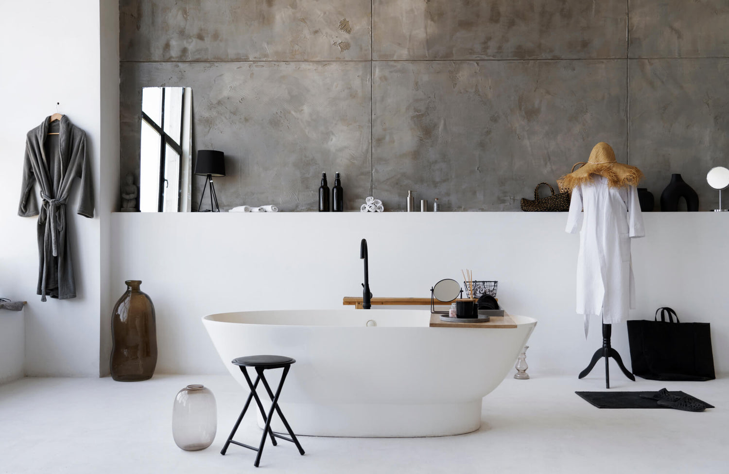 Minimalist bathroom with concrete-look wall tiles behind a freestanding white bathtub, styled with natural accents and softened by warm ambient light from a modern black table lamp.