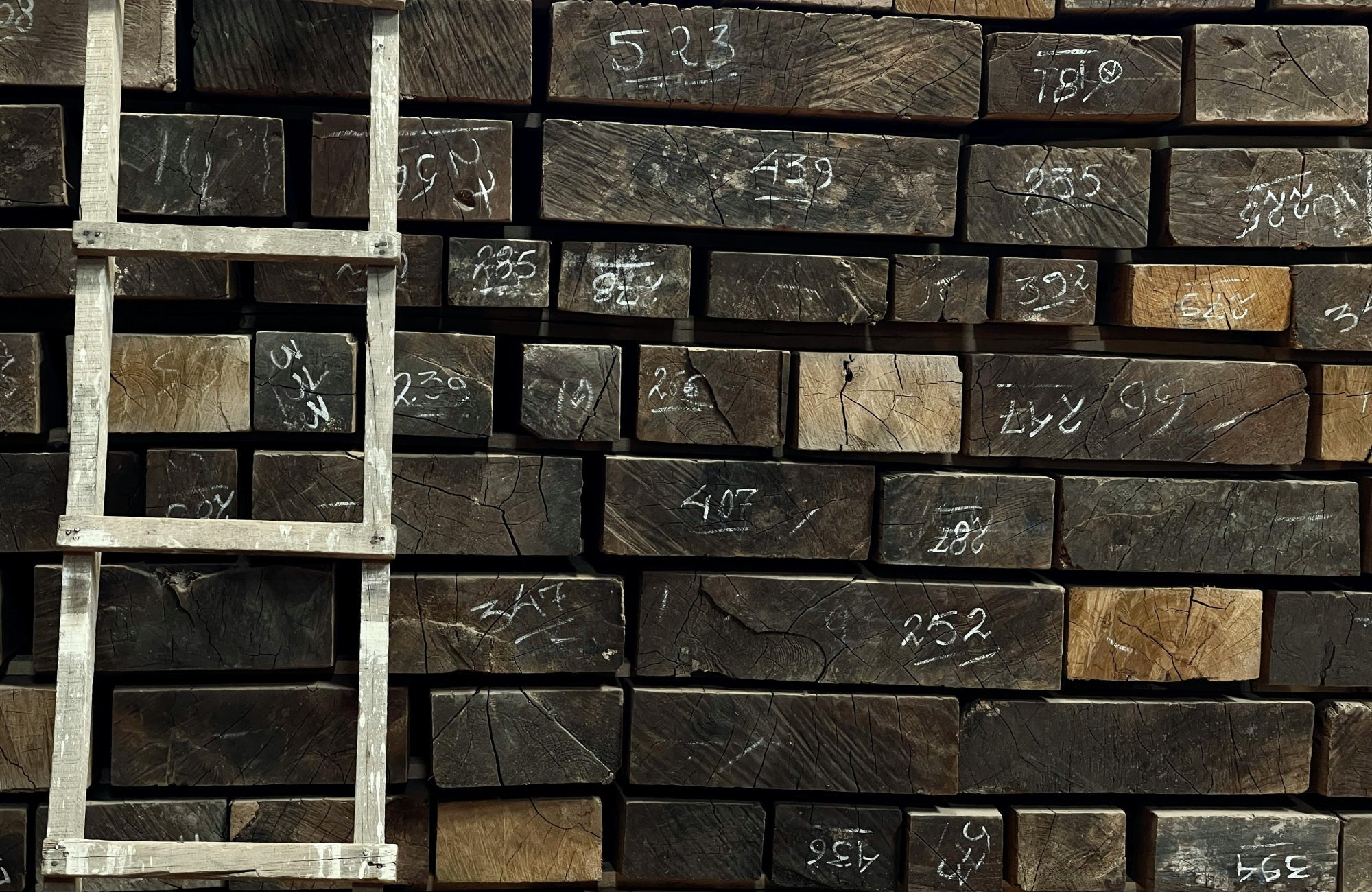 Stacked solid wood beams in a lumber yard marked with chalk measurements, illustrating raw hardwood material used for flooring planks.