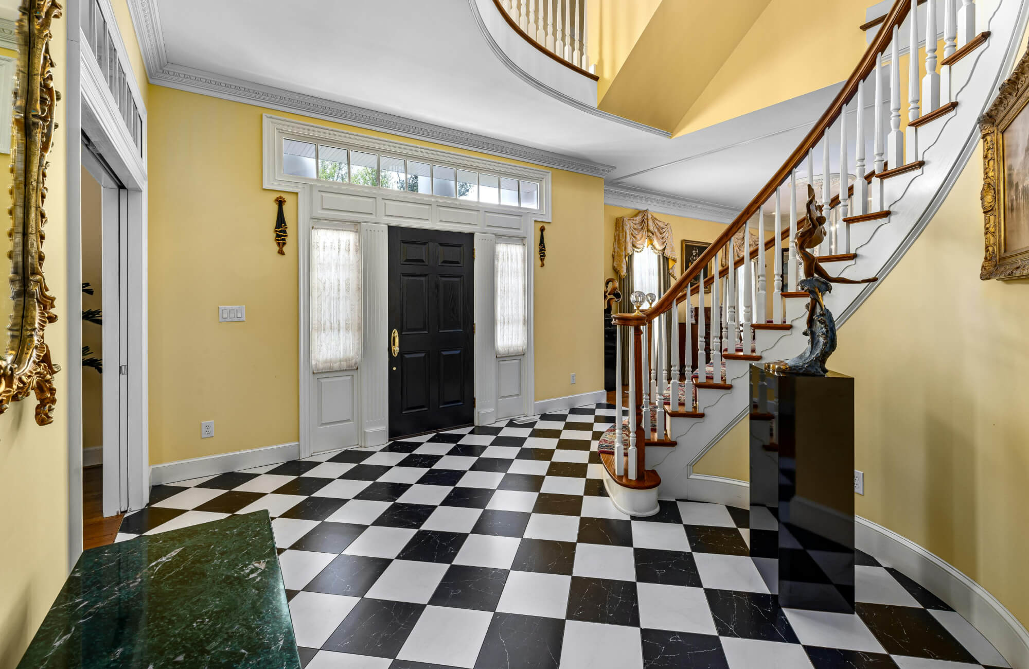 Elegant foyer with black and white checkered marble flooring, a curved staircase with wooden accents, and soft yellow walls.