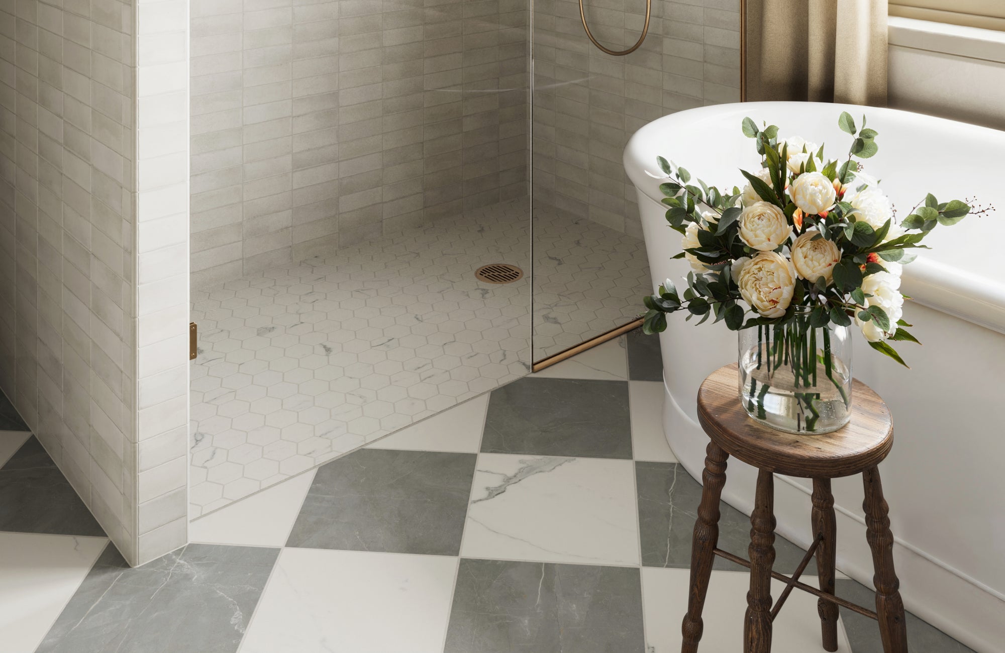 Bathroom with grey and white checkered tiles, a hexagonal-tiled shower accented by brass trim, and a freestanding tub beside a floral arrangement.
