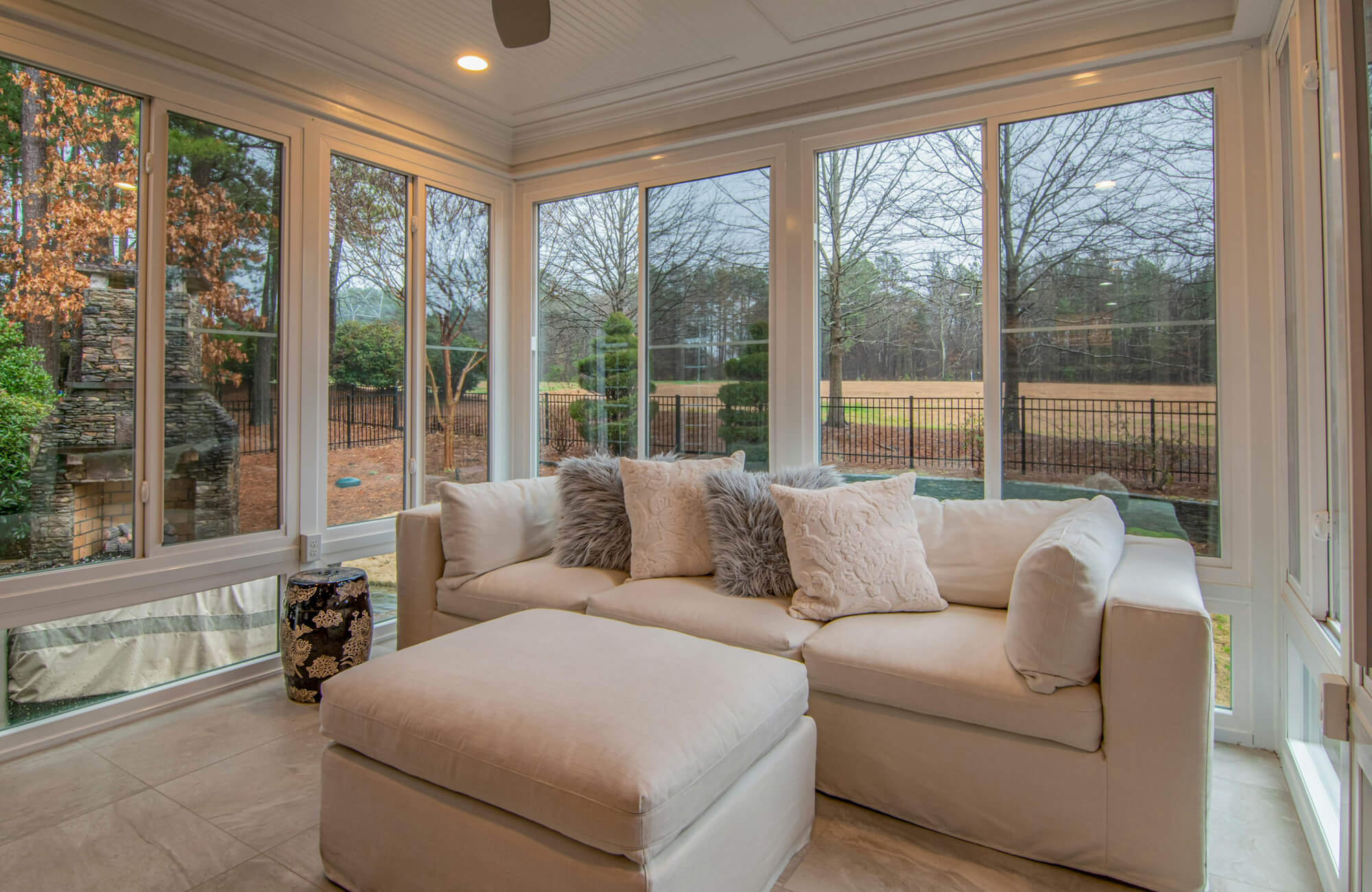 Contemporary sunroom with plush sectional sofa, decorative pillows, large windows, and light stone tile flooring overlooking a serene outdoor landscape.