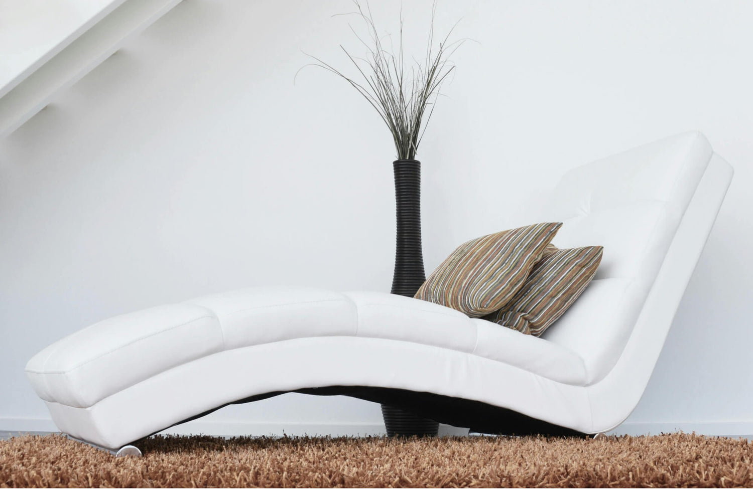 Decorative pillows with striped patterns in warm earth tones placed on a modern white chaise lounge, adding texture and contrast to a minimalist living room setting with a shag rug and tall black vase.