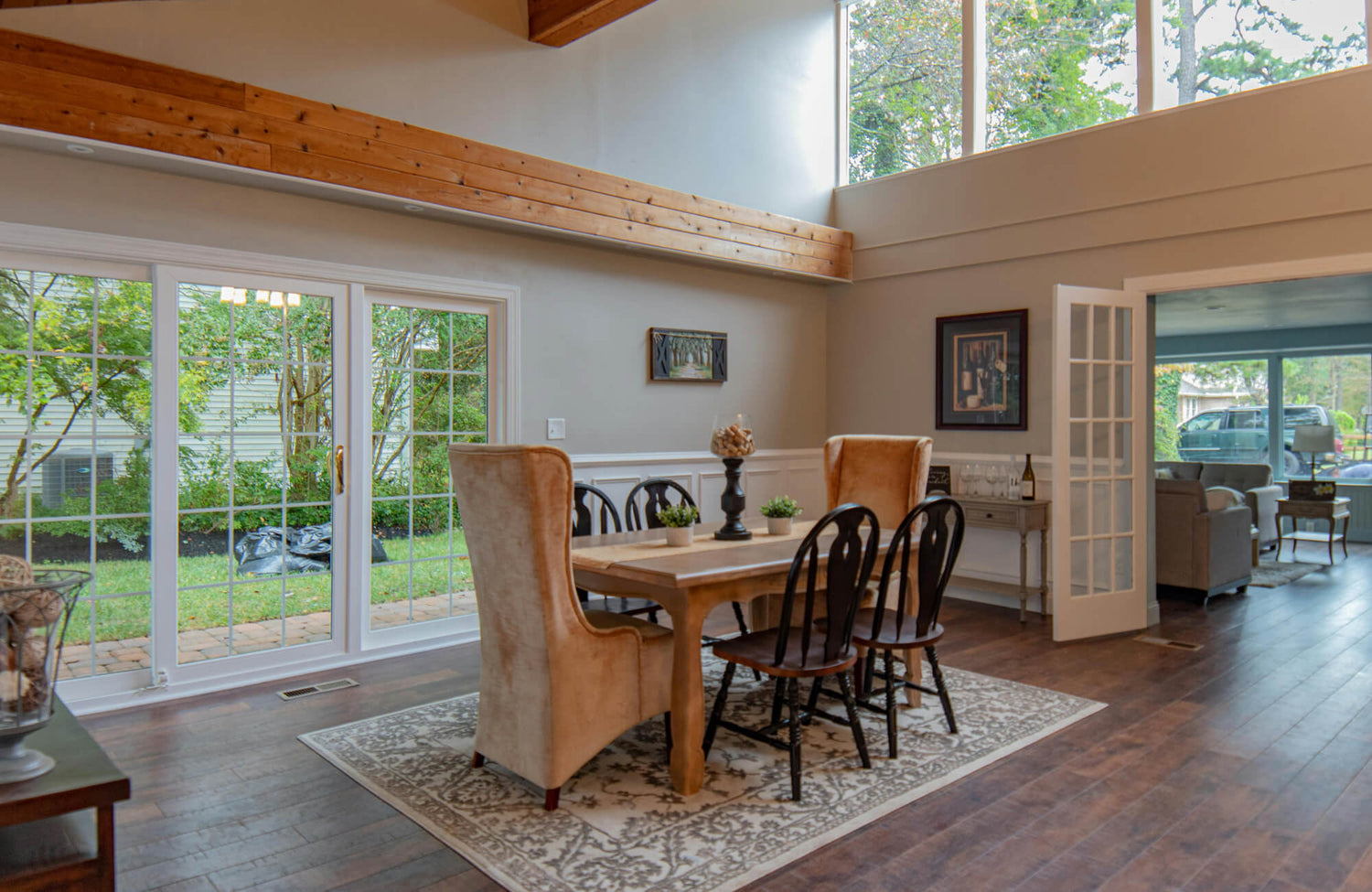 A spacious dining room features a rustic farmhouse table with four chairs on a patterned rug, large windows, and an adjacent living area.