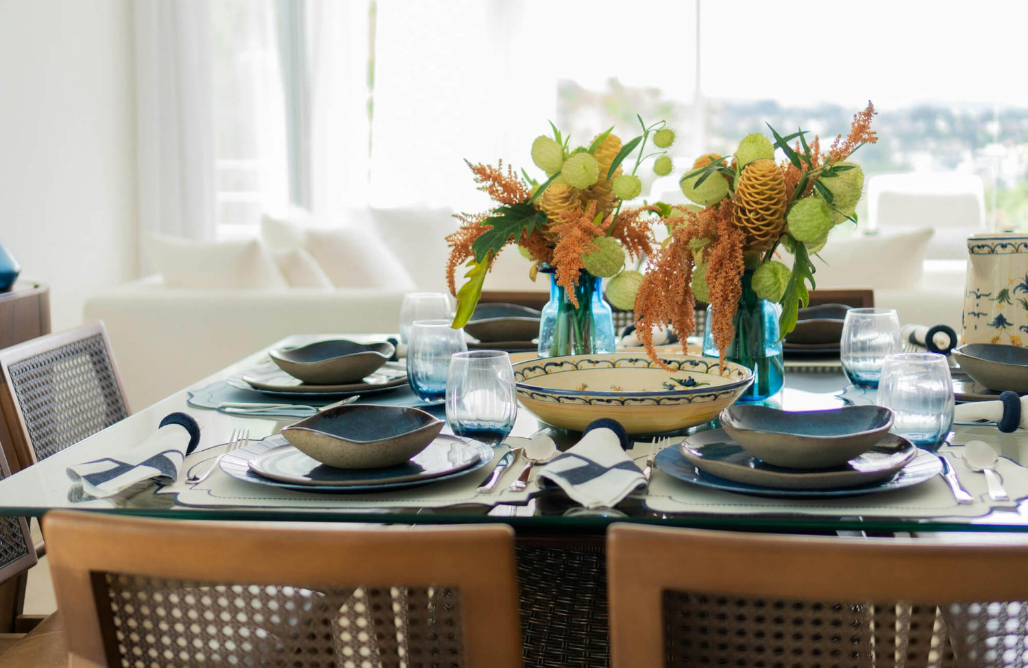 A refined dining setting featuring a sleek glass dining table with a transparent surface that reflects natural light, styled with layered ceramic tableware, blue glass accents, and floral arrangements for a modern atmosphere.