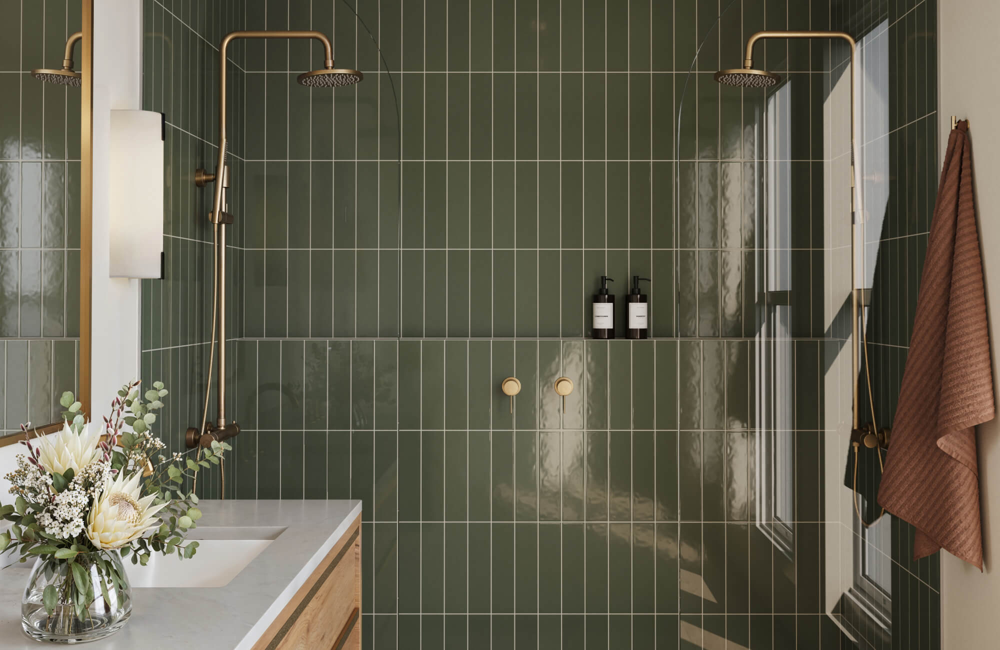 Bathroom with glossy moss green vertical subway tile walls, dual brass shower fixtures, a white vanity top with flowers, and a brown towel hanging beside the shower.