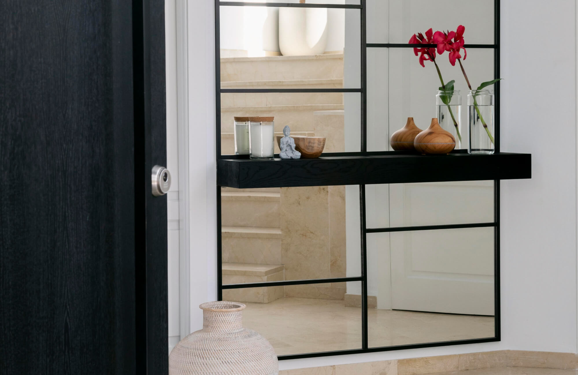 A modern entryway featuring a large floor mirror with black grid-style framing, reflecting the neutral stone staircase and styled with minimalist décor including candles, wooden accents, and fresh florals for a clean, contemporary look.