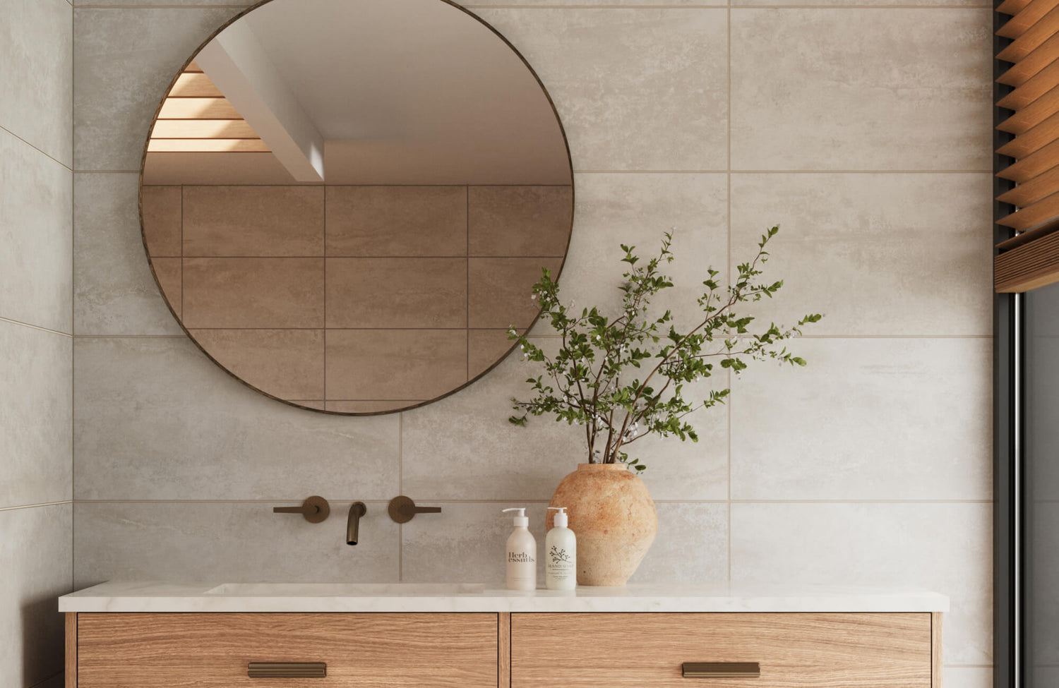 Elegant bathroom wall with large matte beige tiles, featuring a round mirror, minimalist brass fixtures, and a rustic vase with green branches on a wood vanity.