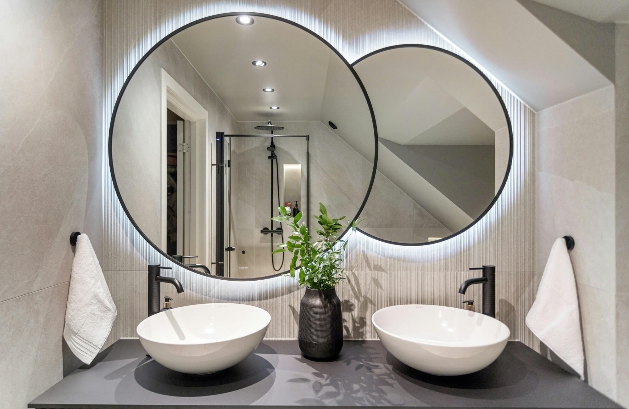 Sleek double vanity with vessel sinks, matte black taps, and LED-lit round mirrors against textured vertical tiles.
