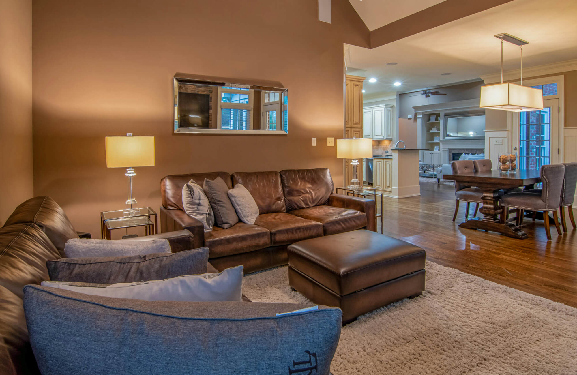 Cozy open-concept living room with leather sofas, gray chairs, and a textured area rug over warm wood floors, next to classic dining area.
