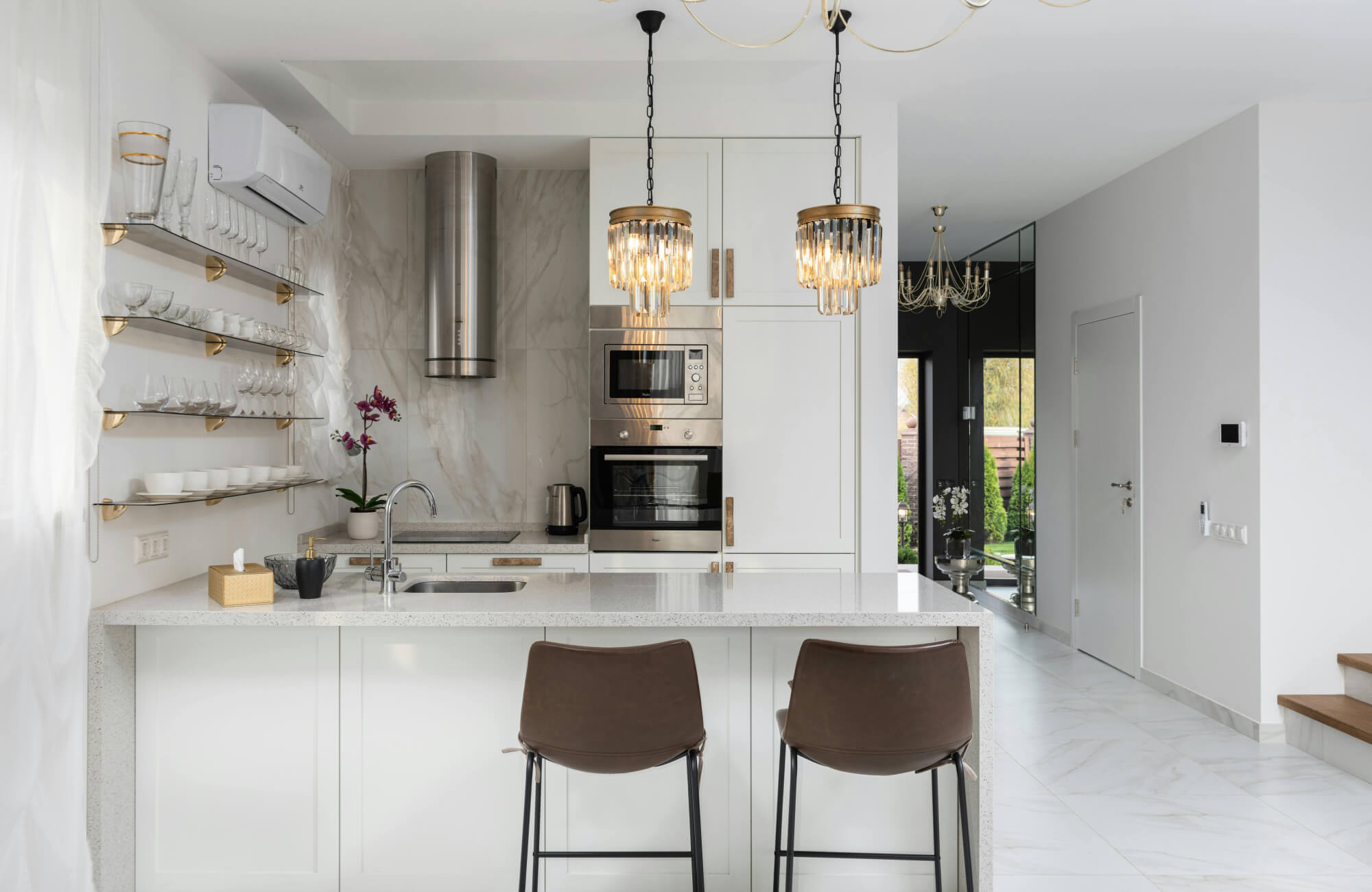 Two modern counter stools with slim black metal legs and curved brown upholstered seats are positioned at a bright white kitchen island, complementing the clean cabinetry, marble-look backsplash, and warm brass lighting.