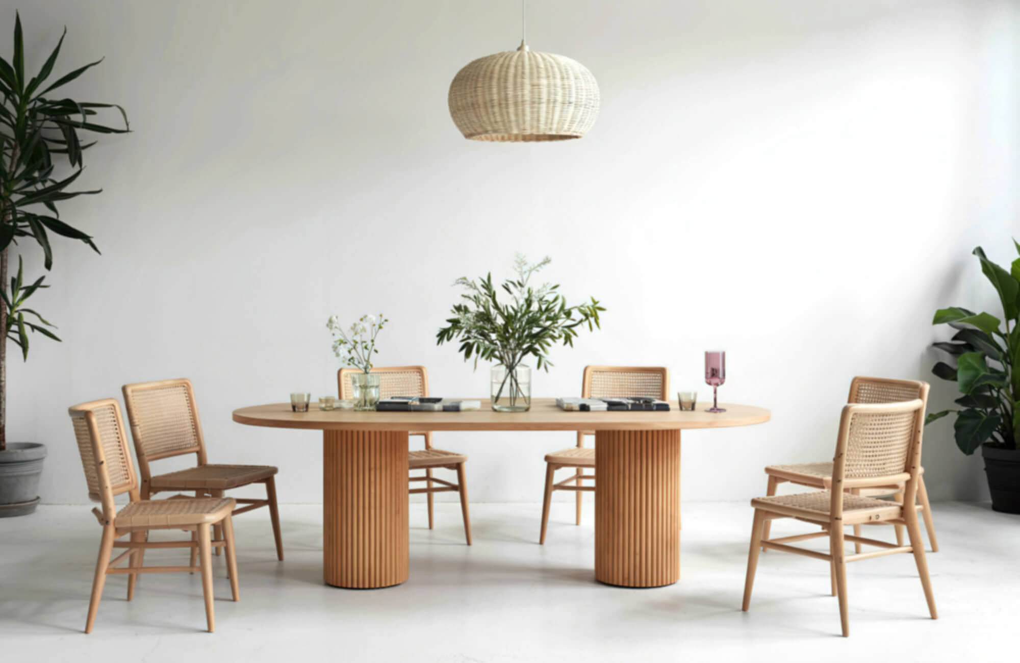 Natural wood oval dining table with woven chairs, greenery accents, and soft modern styling in a bright, minimalist room.