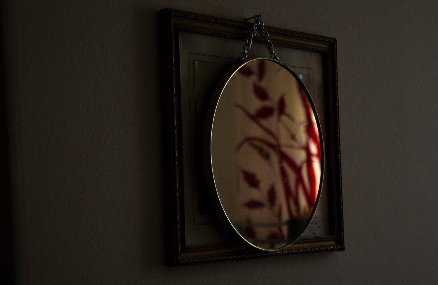 Oval wall mirror with a slim metal frame hung against a dark wall, reflecting soft shadows and creating visual depth that helps narrow spaces feel calmer and less rigid.