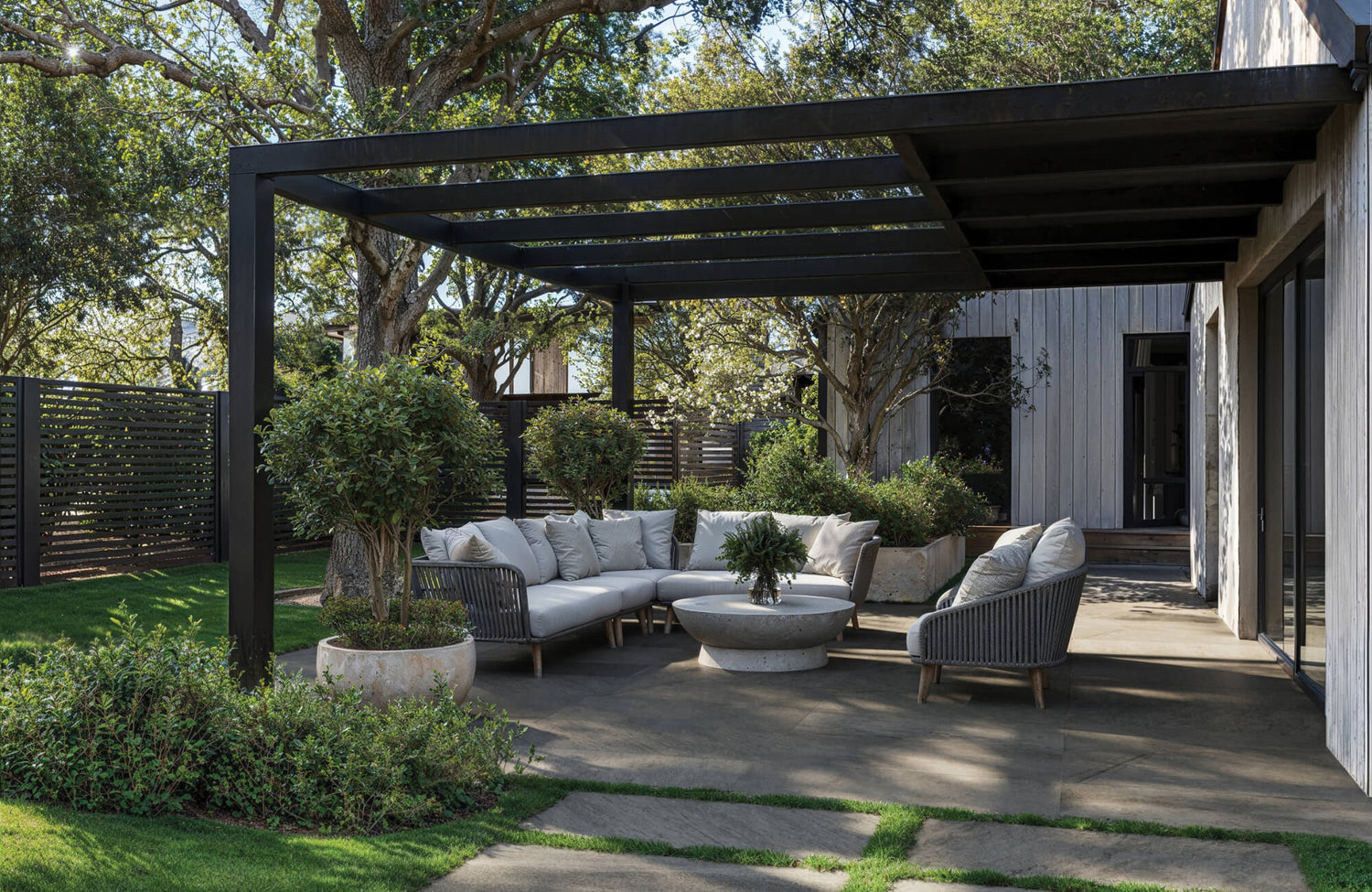 Modern outdoor patio with porcelain pavers, a black pergola, cushioned sectional seating, and surrounding greenery in a garden setting.