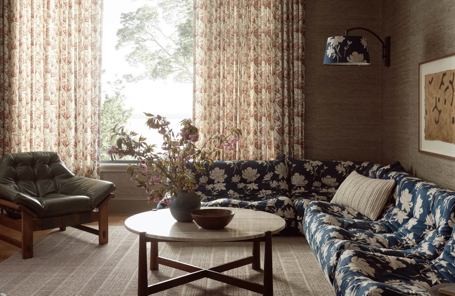 Cozy living room with a blue floral sectional, patterned rug, botanical curtains, and a round wood coffee table with floral arrangement.