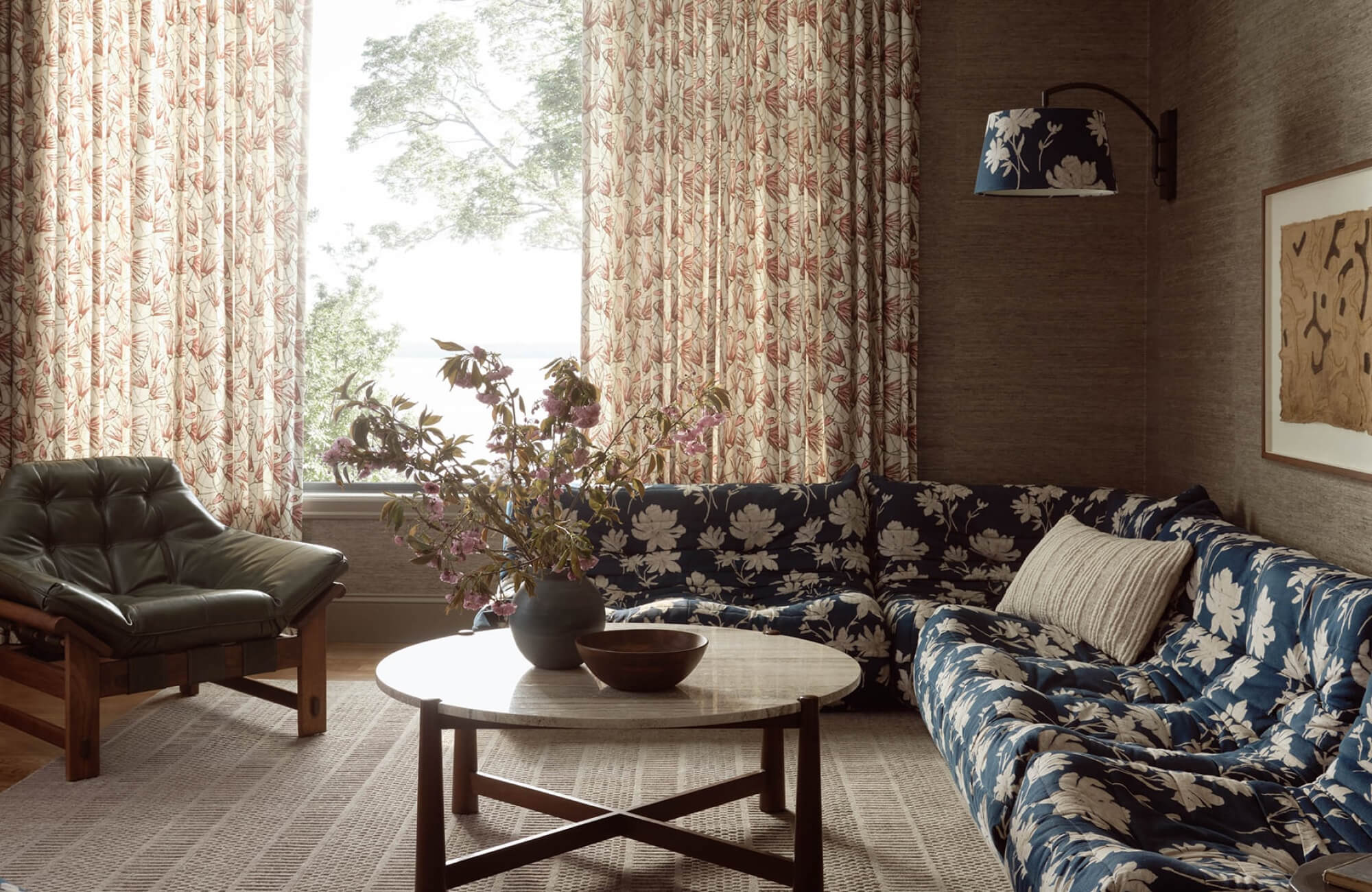 Cozy living room with a blue floral sectional, patterned rug, botanical curtains, and a round wood coffee table with floral arrangement.