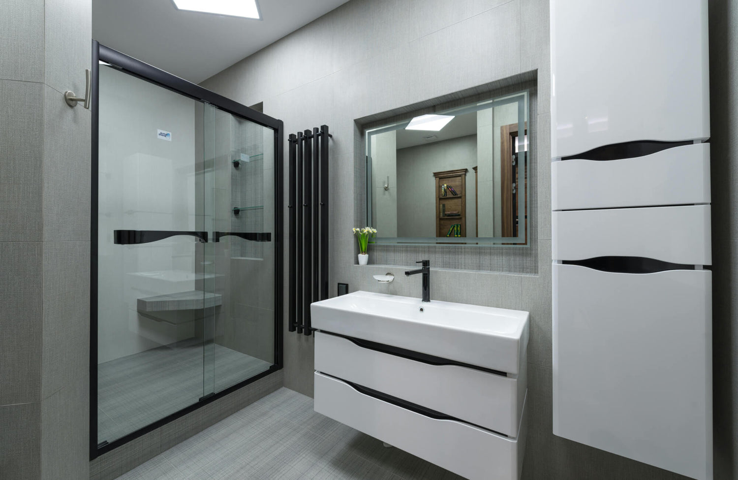 A clean, contemporary bathroom featuring a sliding glass shower door, white floating vanity, black faucet, and a square mirror framed by smooth gray tile.