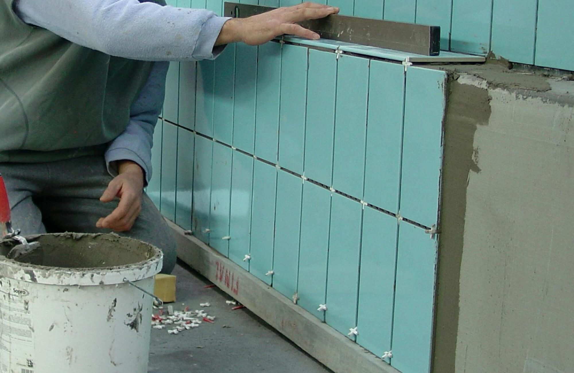 Skilled worker aligning glossy aqua-blue subway tiles on a wall, with precision tools and an adhesive bucket nearby, creating a sleek modern finish.