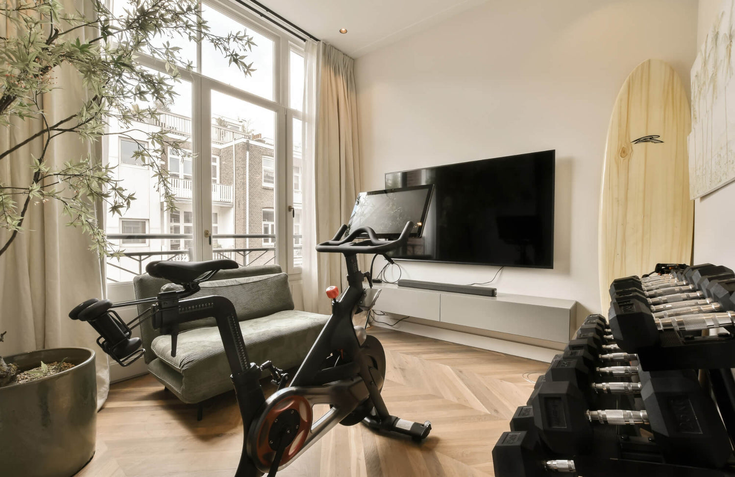 A modern home gym with a stationary bike, dumbbells, and a TV, featuring herringbone wood-look tile flooring and large windows for natural light.