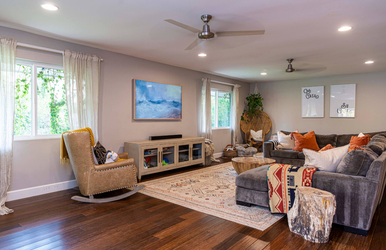 Bright, cozy living room with hardwood floors, a patterned wool rug, neutral furniture, and large windows with sheer white curtains.