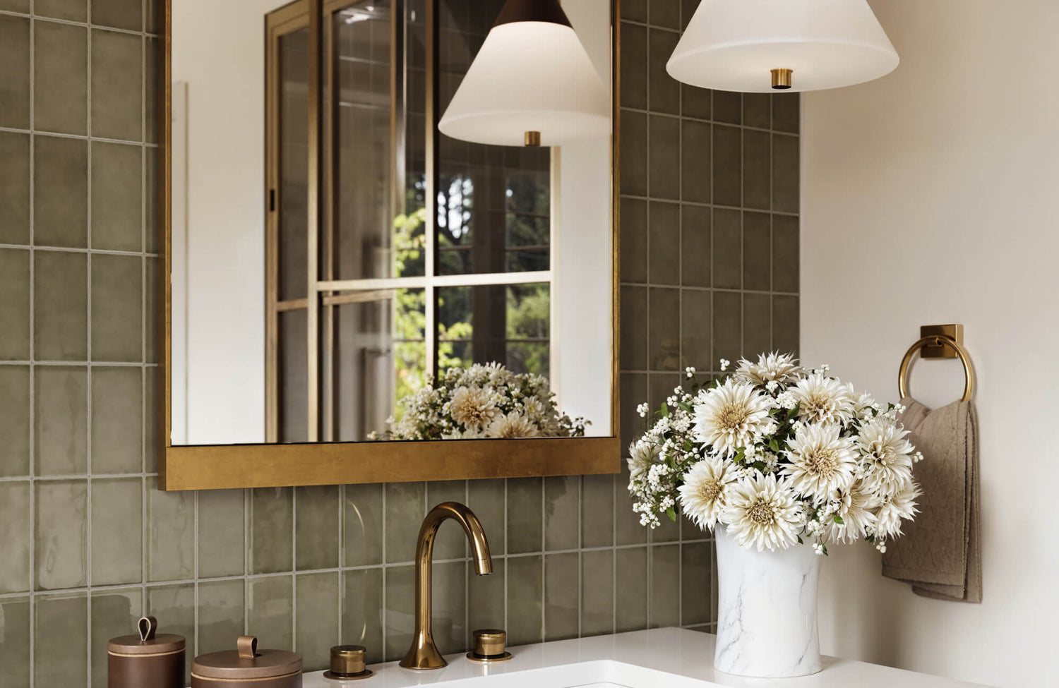 Bathroom vanity with glossy olive green square wall tiles, a gold-framed mirror, and a marble vase of white flowers beside a modern brass faucet.