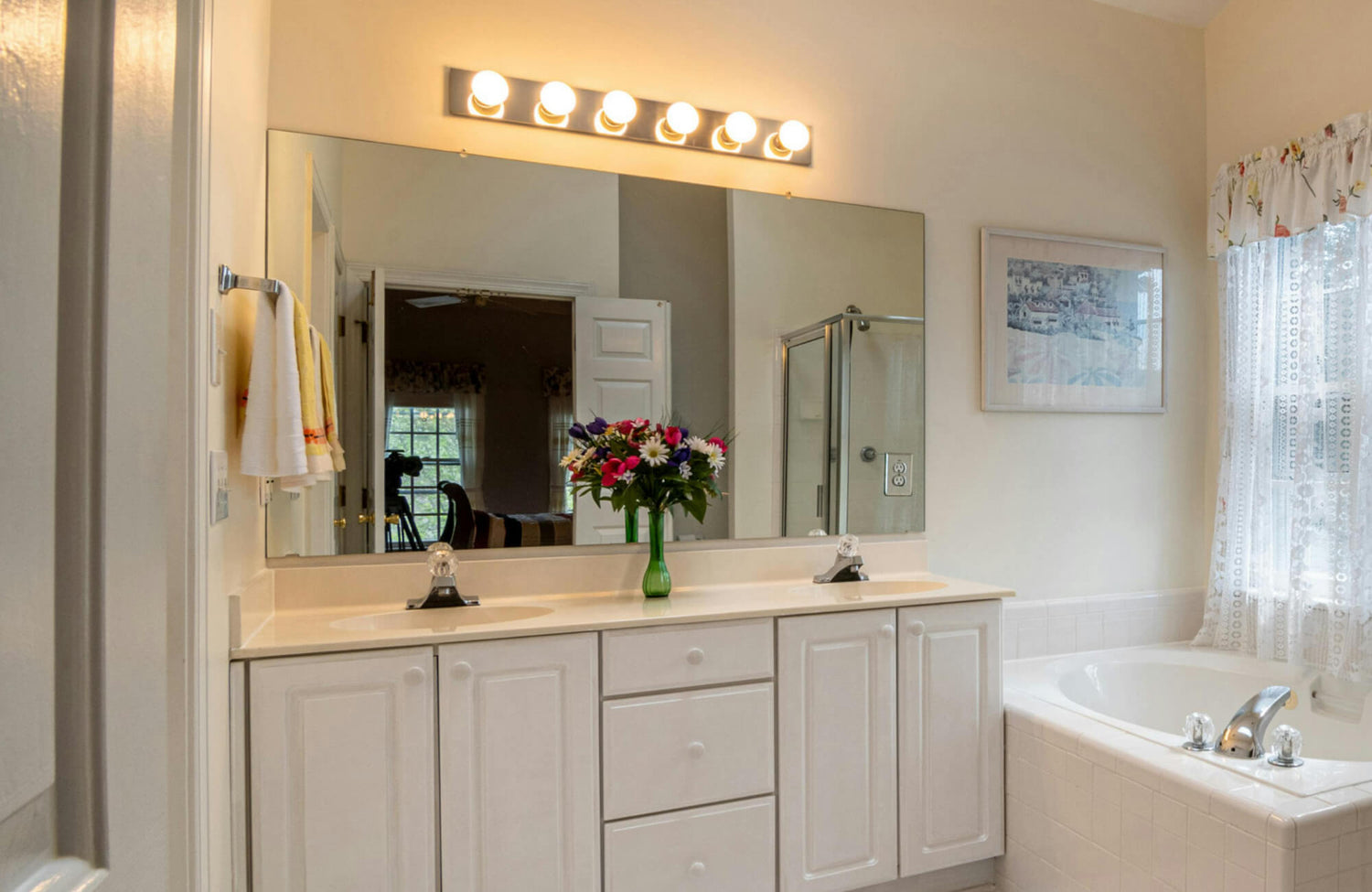 Bright bathroom vanity lighting with a row of globe bulbs above a large mirror, enhancing the clean white double-sink vanity and cozy tub space.