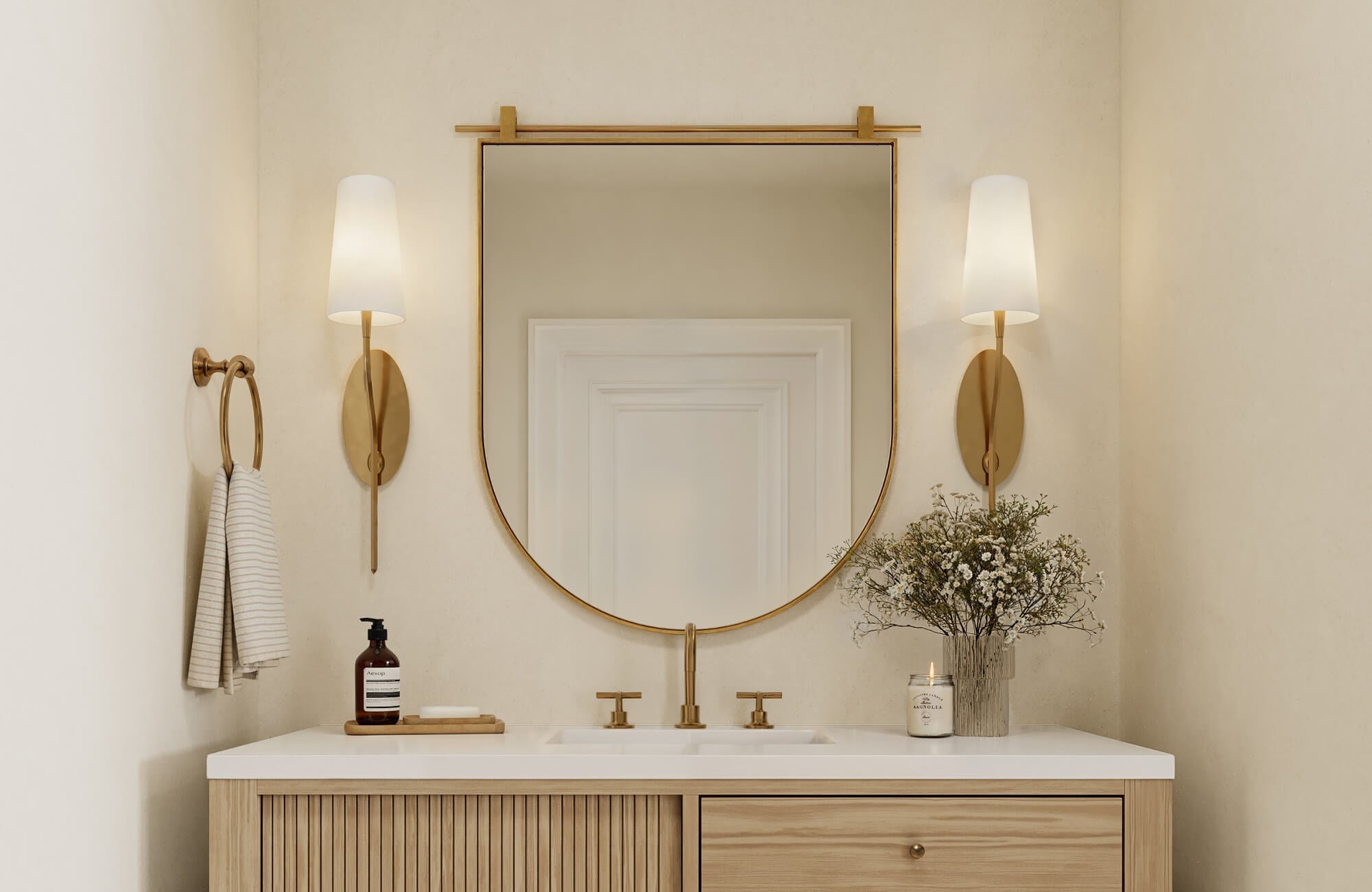 Bathroom vanity with light wood cabinet, gold-framed mirror, matching gold sconces, and a white countertop decorated with flowers and a candle.