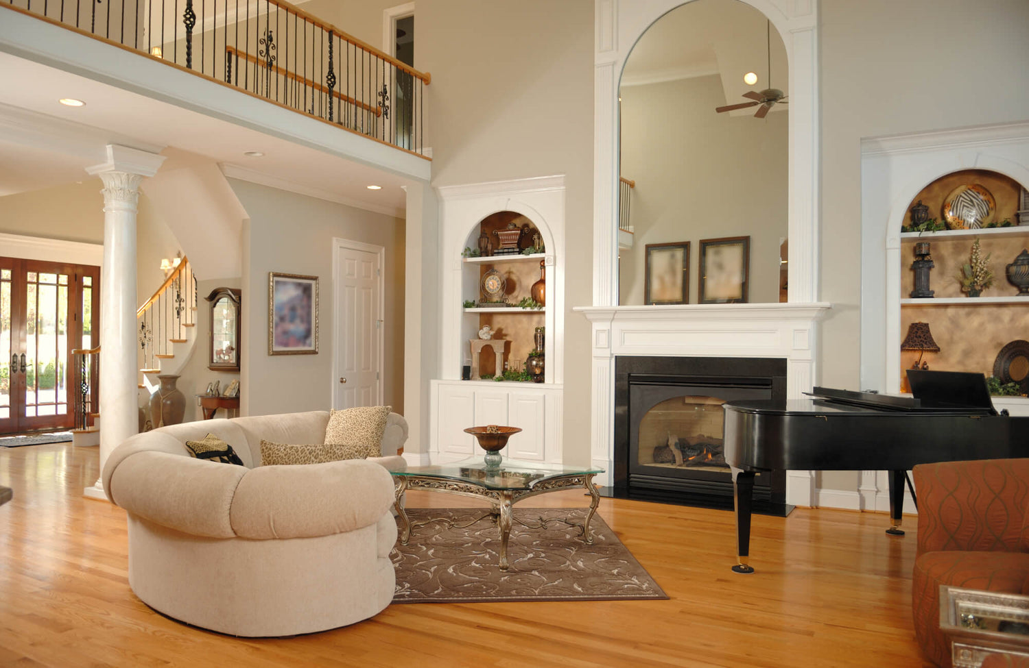 Elegant living room with a beige circular sofa, brown patterned rug, glass coffee table, grand piano, and built-in white shelves.