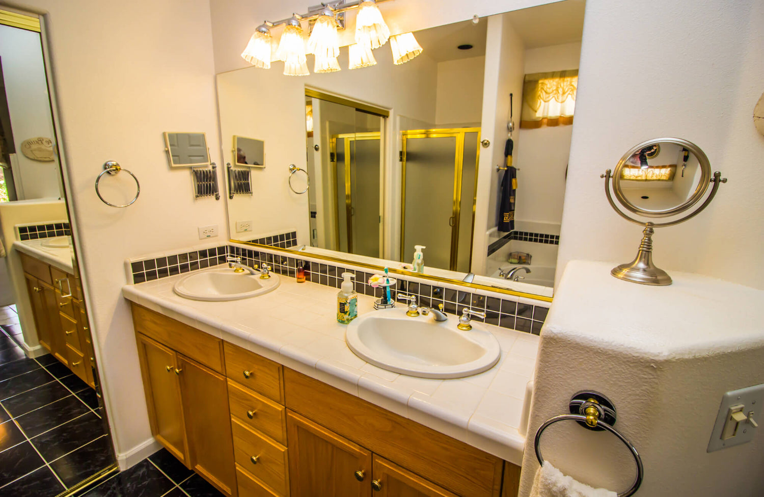 Double vanity bathroom with wood cabinetry, dual sinks, large wall mirror, and black tile accents under warm overhead lighting.