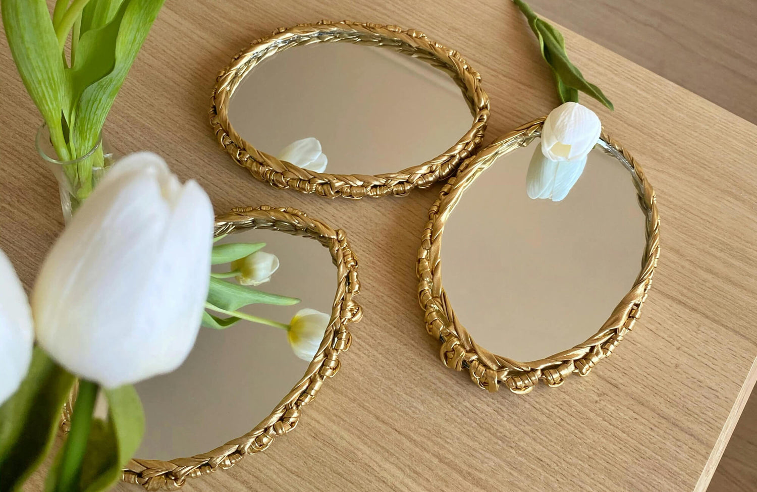 Decorative gold-framed mirrors with braided detailing displayed on a wooden tabletop, paired with fresh white flowers for a warm, modern vignette.