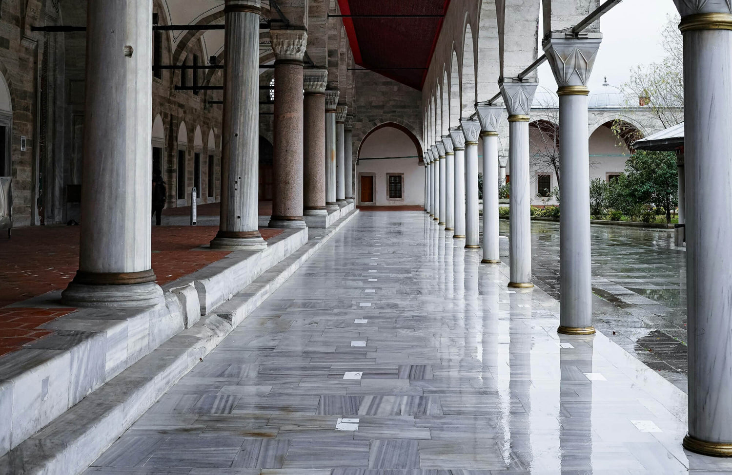 Elegant covered walkway with polished marble flooring, grand columns with gold accents, and arched openings overlooking a courtyard.