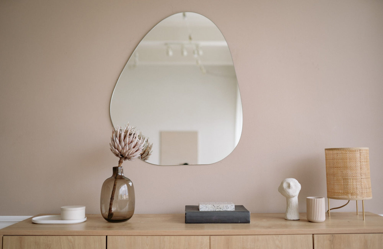 Organic-shaped wall mirror above a wooden console with minimalist decor, including a vase, sculpture, books, and a rattan lamp.