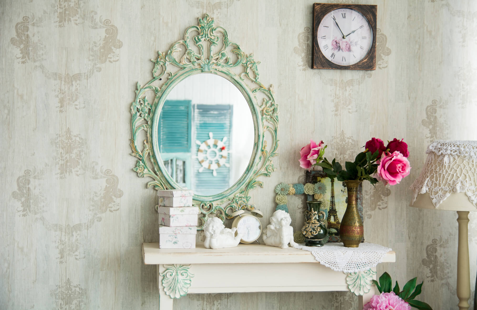 Vintage-style vanity with ornate oval mirror, floral decor, antique clock, and lace lamp against patterned wallpaper backdrop.
