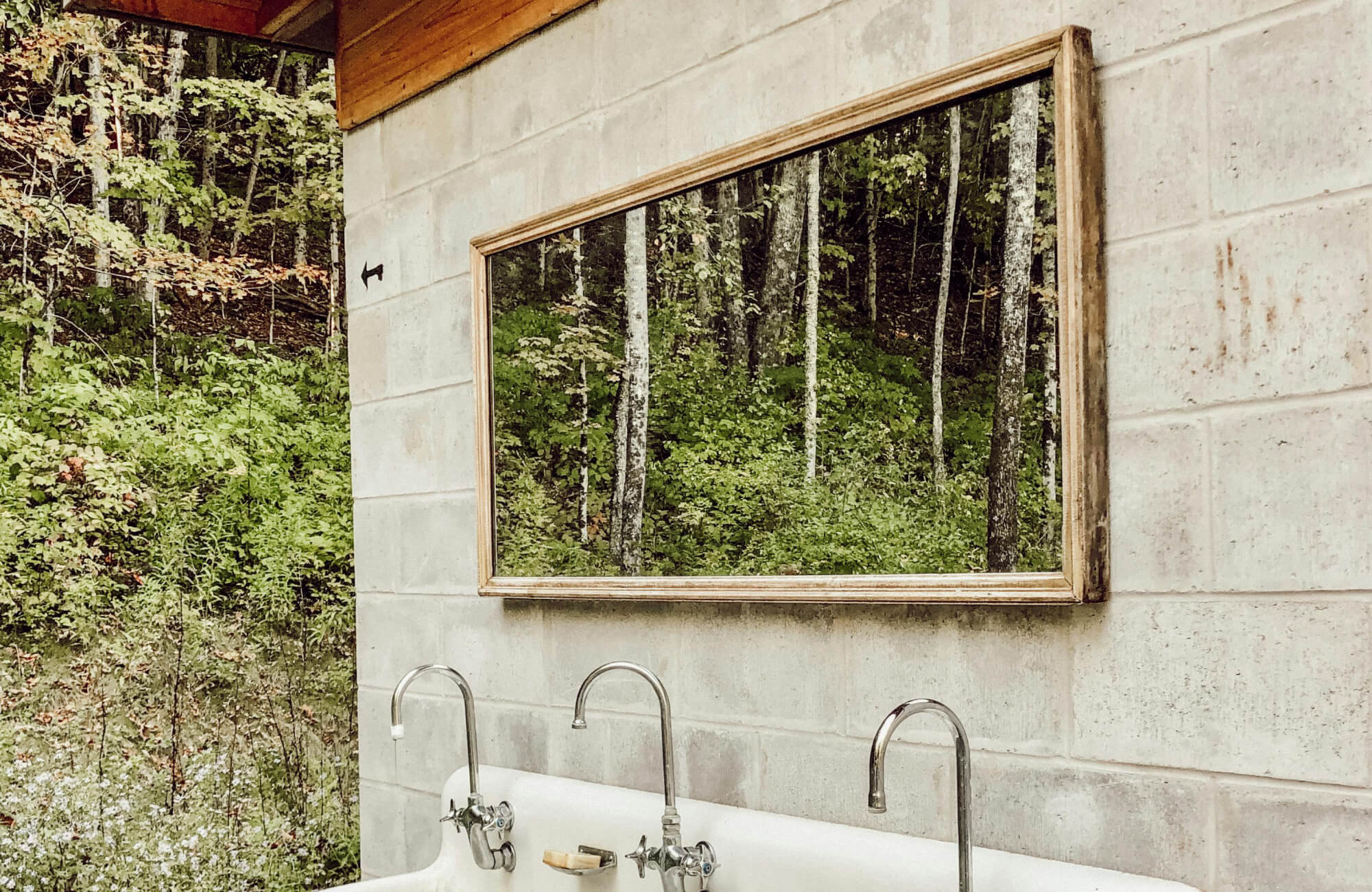 Weathered wood-framed mirror used in a semi-outdoor wash area, doubling the surrounding woodland view to create depth and connection with nature.
