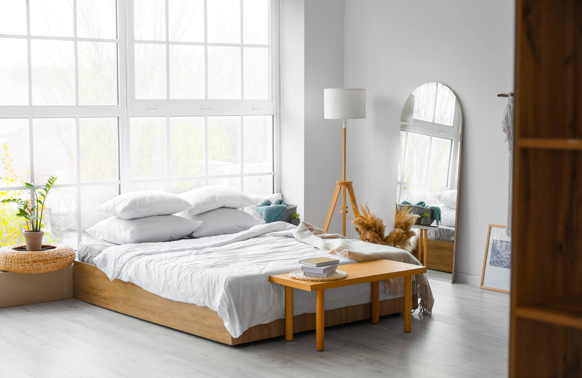 Modern bedroom with large windows, a low wooden bed, and an arched floor mirror reflecting natural light and soft, neutral decor.