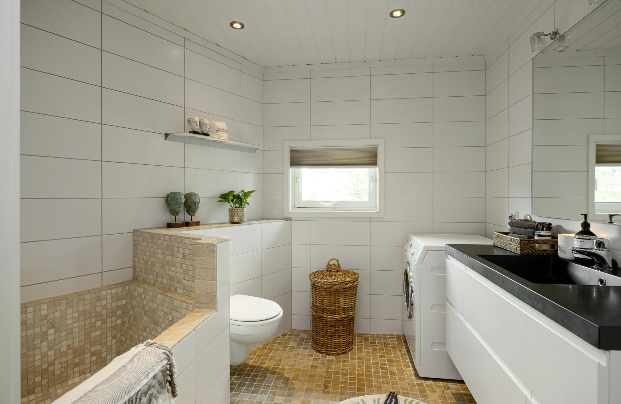 Modern bathroom with beige mosaic tile flooring and tub surround, large white wall tiles, black countertop, and built-in laundry unit.