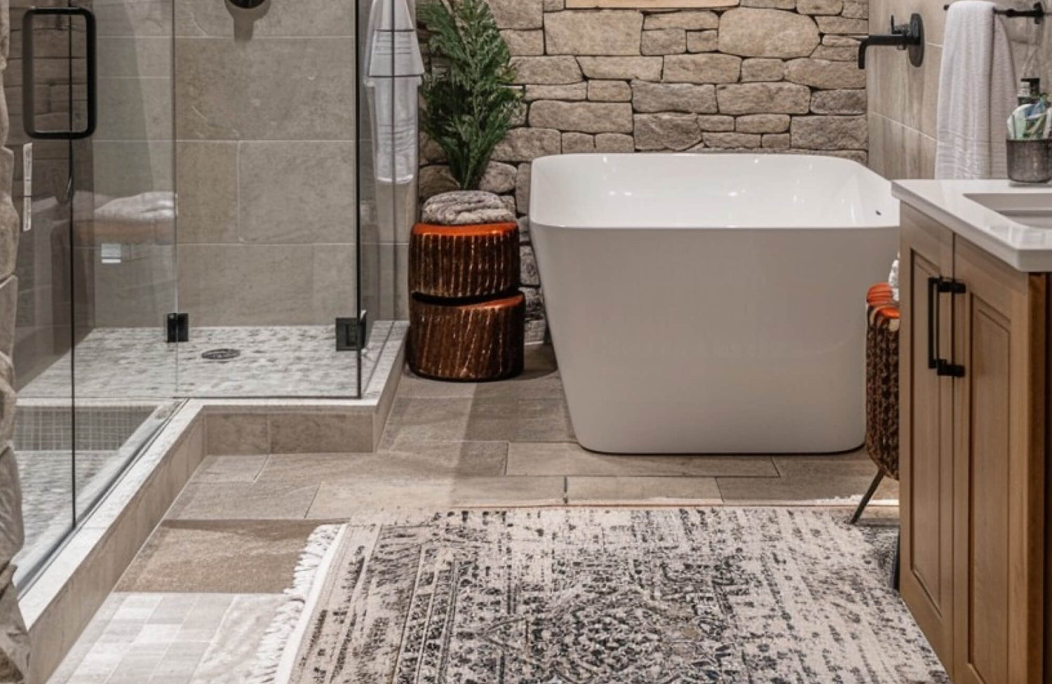 Luxurious bathroom with a freestanding tub, glass-enclosed shower, and textured stone wall, featuring warm-toned floor tiles with polypropylene rug.