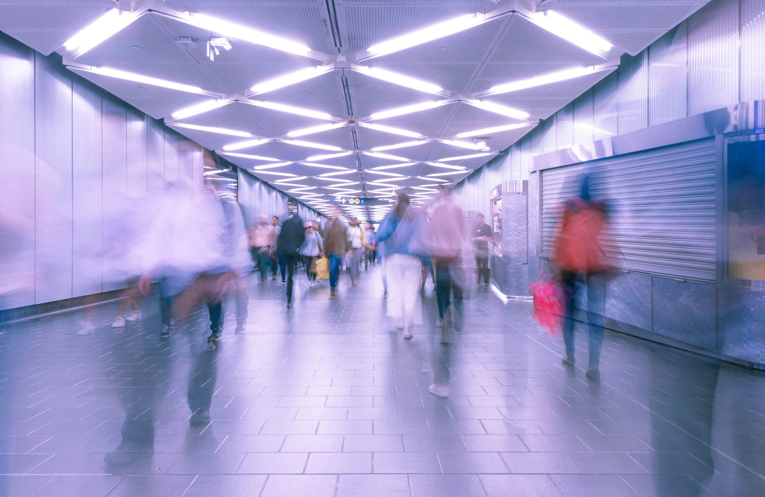 People walking through a brightly lit subway corridor with dark floor tiles and geometric ceiling lights, creating a motion blur effect.