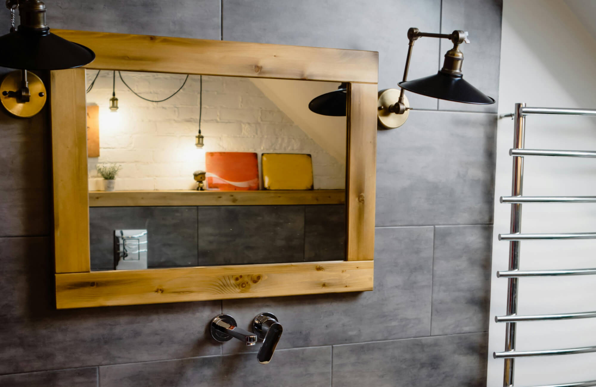 Bathroom with a rectangular wood-framed mirror, gray tiled wall, and industrial-style black sconces beside a chrome towel radiator.