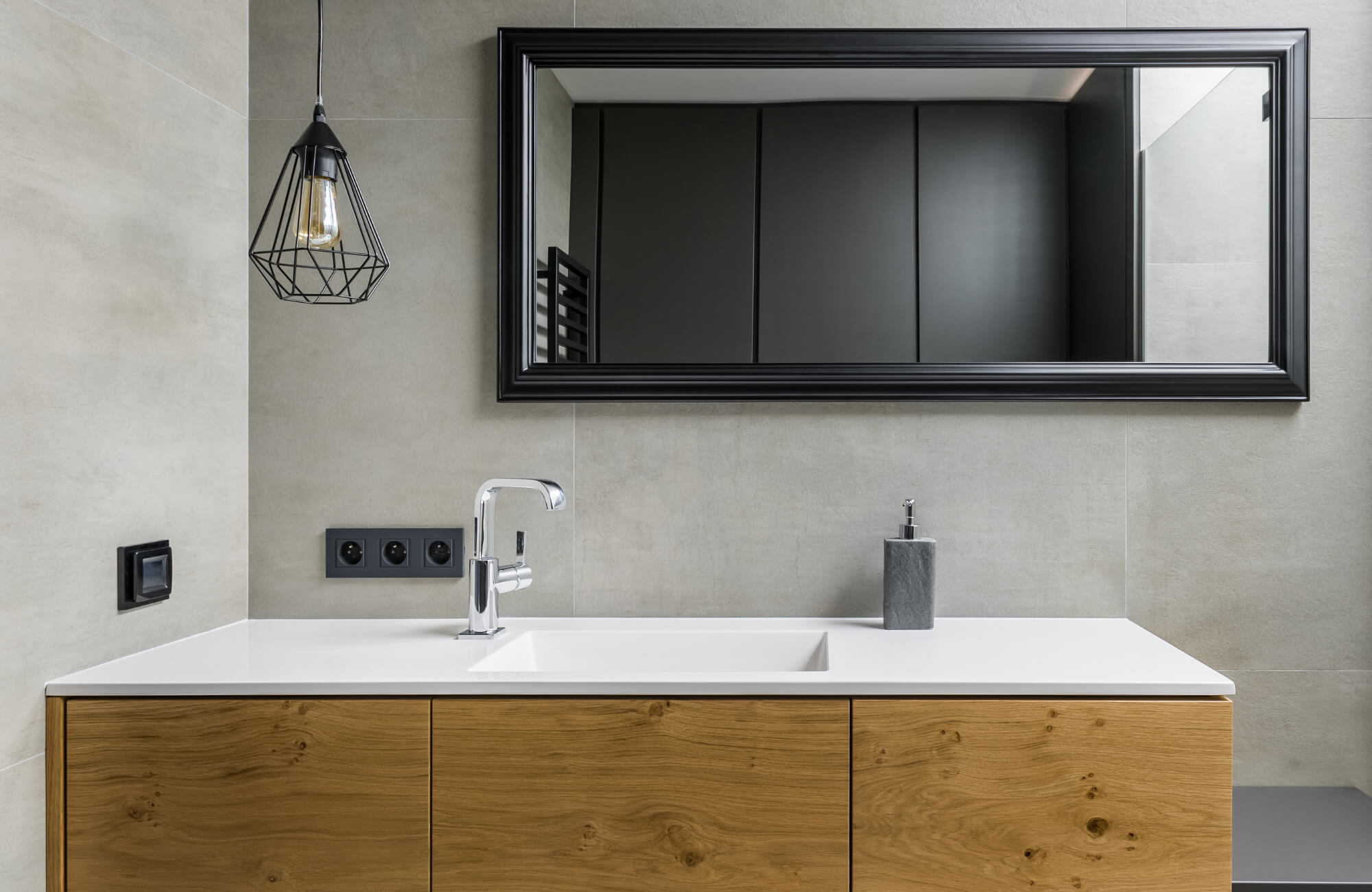 Modern bathroom with a black pendant light, chrome faucet, wood vanity, and large black-framed mirror on neutral tile walls.