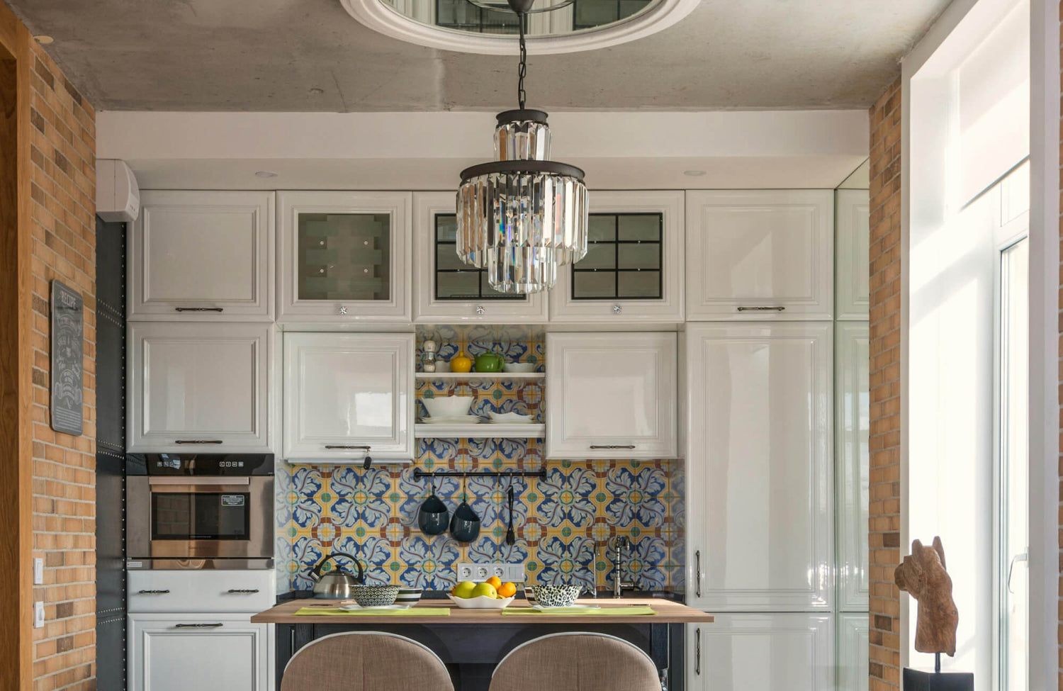 A crystal chandelier adds a touch of elegance above a vibrant Spanish-style backsplash and glossy white kitchen cabinets.