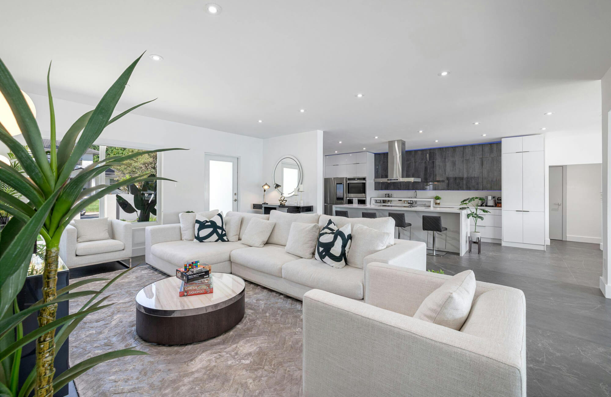 Modern open-concept living room with neutral sectional sofa and patterned pillows, styled with plants, marble coffee table, and sleek kitchen backdrop.