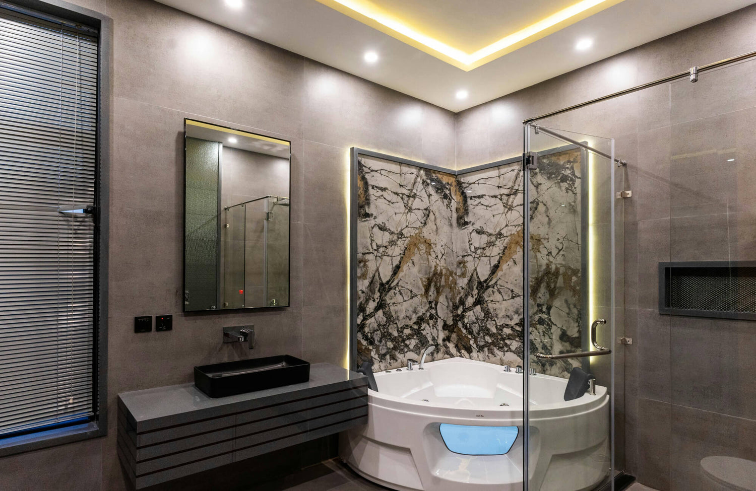 Modern bathroom with large format gray tiles, glass shower enclosure, marble accent wall, and built-in lighting around a white tub.
