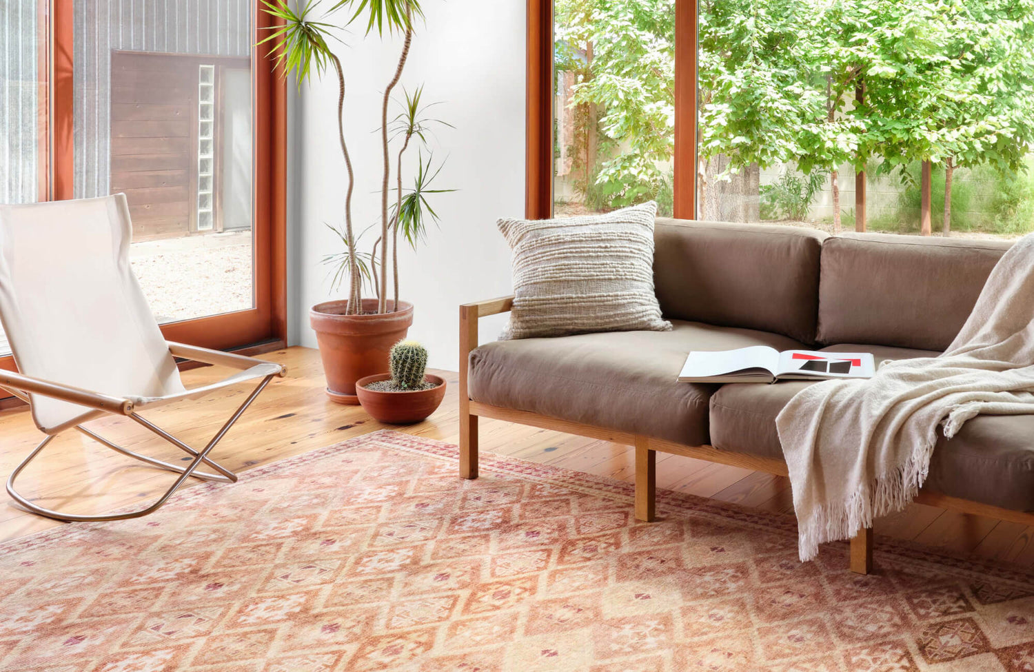 Sunlit living room with a tan sofa, potted plants, and a patterned rug over wood floors, featuring large windows and lush outdoor views.