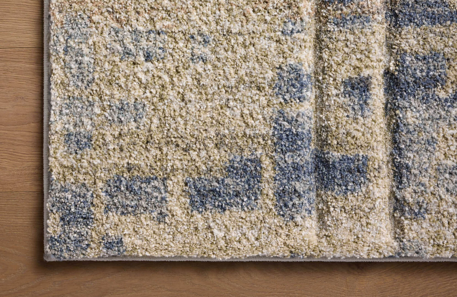 Close-up of a thick, textured rug corner on a warm wood floor, showing an abstract beige, taupe, and muted blue pattern with a bound edge.