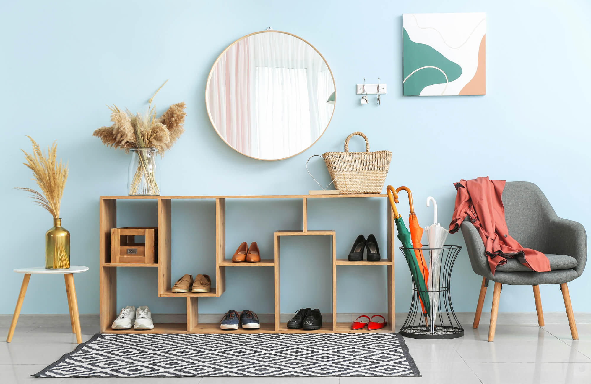 Round mirror above wooden entryway storage with shoes, wicker bag, umbrella stand, chair, and decor against light blue wall.