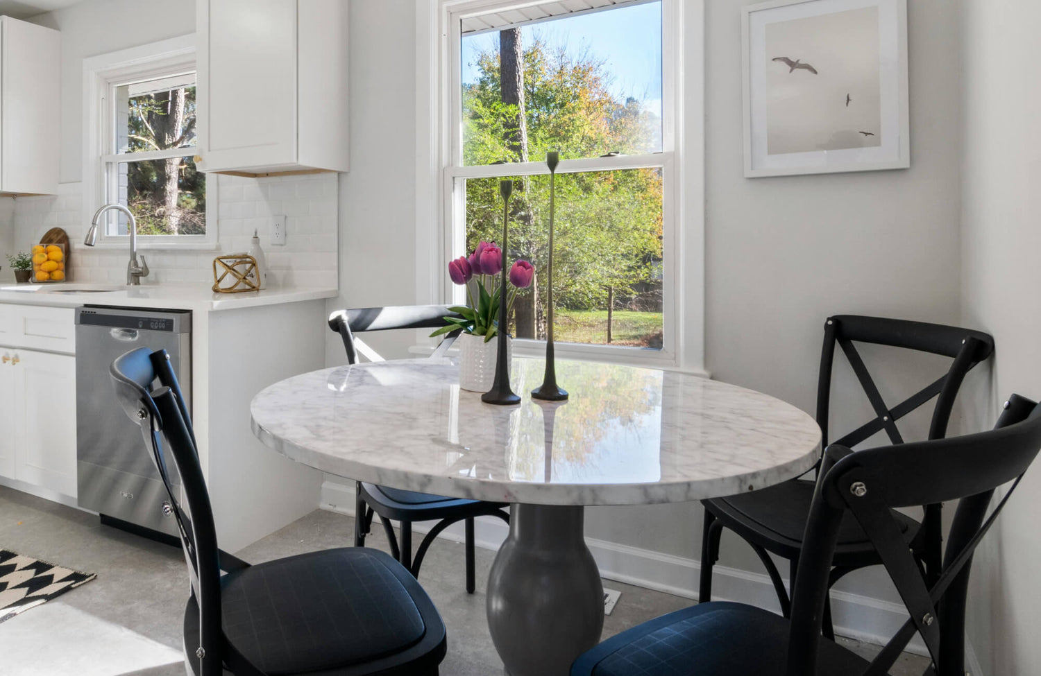 Bright breakfast nook with a round marble-top table, black cross-back chairs, and a vase of tulips by a sunlit window.