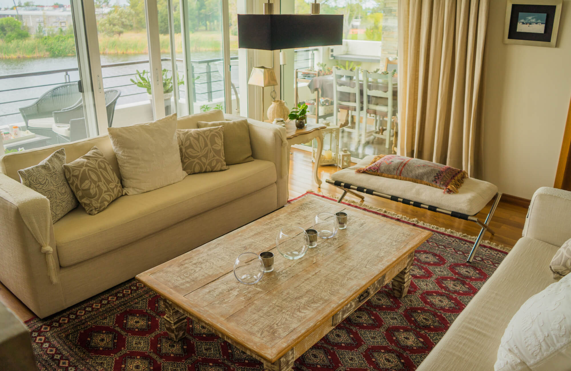 A bright, inviting living room features neutral upholstered sofas and a rustic wood coffee table layered over a richly patterned vintage rug in deep red and earth tones, adding warmth, contrast, and timeless character.