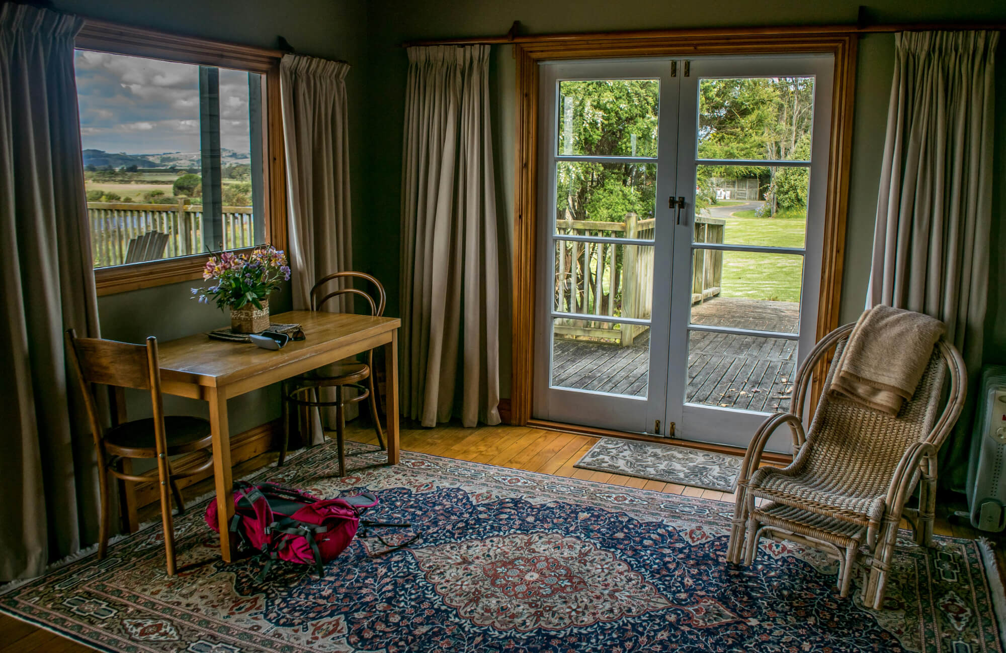 Classic navy and red Persian rug adds charm to a cozy country room with wood furniture, wicker chair, and garden views.