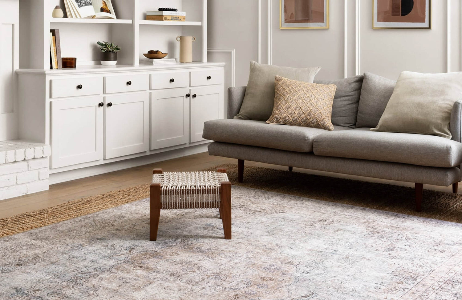 A cozy living room with a neutral palette, featuring a soft gray sofa, woven ottoman, and a vintage-style area rug over warm wood flooring.