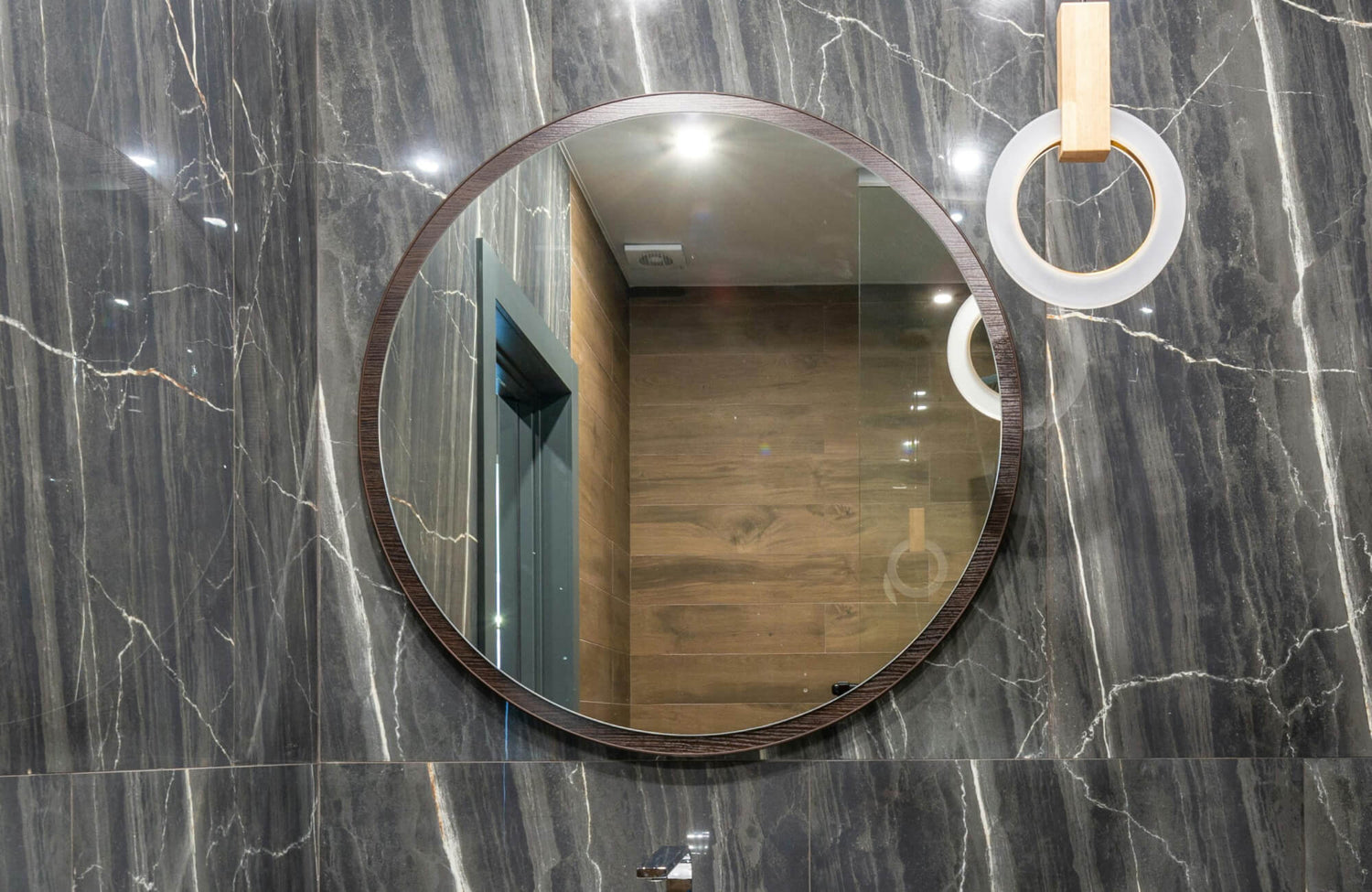 Round bathroom mirror with a dark wood frame mounted on polished gray marble walls, reflecting warm-toned wood paneling.